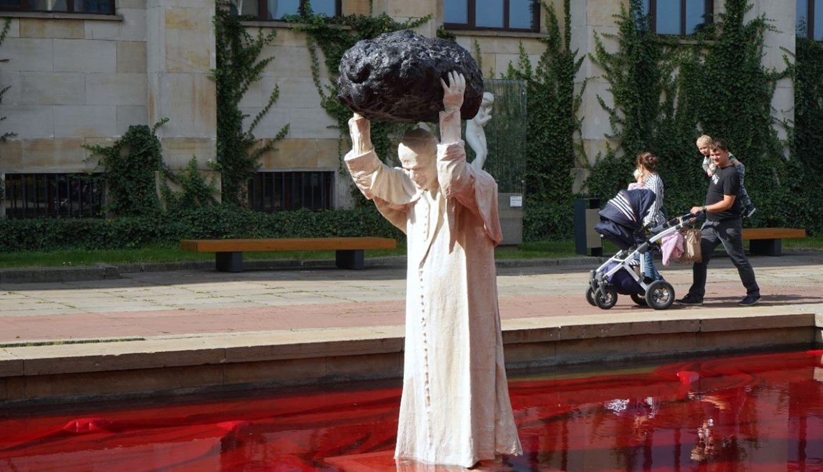 منحوتة أمام المتحف الوطني في وارسو، للنحات البولوني جيرزي كالينا، تمثل البابا الراحل يوحنا بولس الثاني وهو يحمل نيزكًا فوق رأسه، واقفا في بركة من الماء الأحمر رمزا للدم (24 أيلول 2020، أ ف ب).  