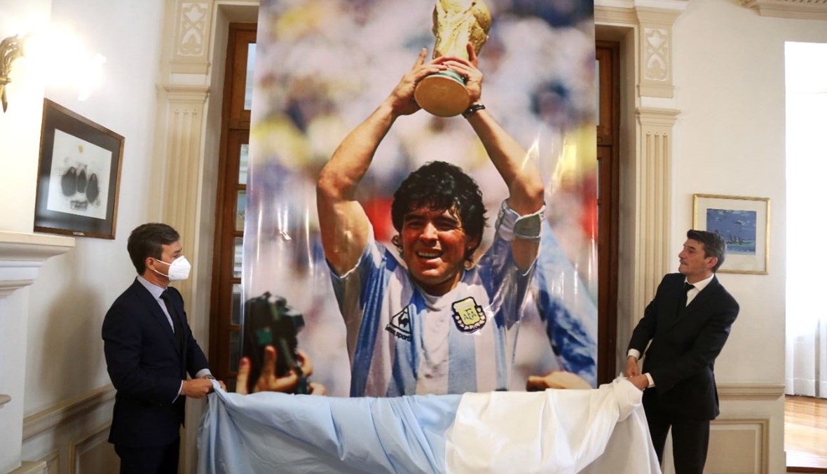 القائم بأعمال السفارة الأرجنتينية في بوليفيا دييغو ألونزو غارسيس (إلى اليمين) يكشف صورة لمارادونا خلال تكريم له في مقر السفارة في لاباز (27 ت2 2020، أ ف ب). 