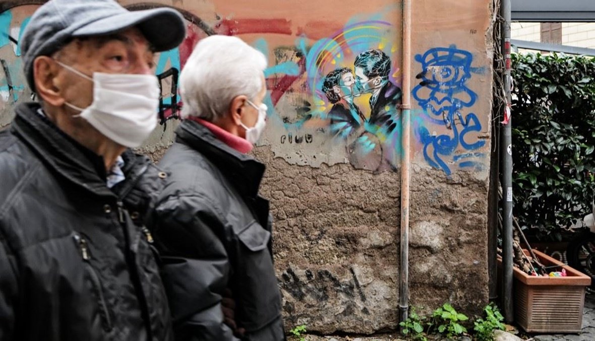 رجلان يمران بشارع على جدرانه رسم يصور قبلة بين رجل وأمرأة وقد ارتديا كمامات للوقاية من كورونا في روما أمس.   (أ ف ب) 